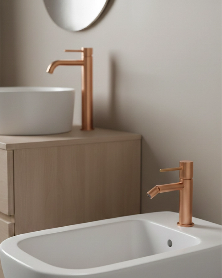 Coppia rubinetti lavabo alto e bidet senza scarico oro rosa in ottone e satinato ambientato su piano bagno con accessori...
