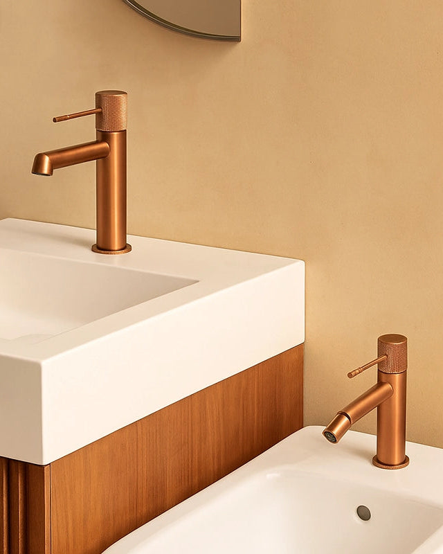 Coppia rubinetti lavabo e bidet senza scarico oro rosa in ottone e satinato ambientato su piano bagno con accessori...